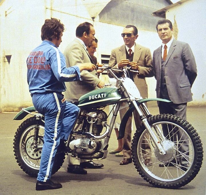 The last iteration of the original Ducati Scrambler family was the 450 R/T, here in motocross trim for testing. The Doctor of Desmology himself, Fabio Taglioni (wearing shades),discusses the machine with the corporate chiefs. Longtime development rider/racer, Franco Farne, wears NCR race shop work clothes with the slogan "Scuderia Speedy Gonzales." The 450 had a desmo valvetrain and weighed 285 lbs wet.