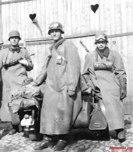 Motorcycle Gear circa 1941: The distinctive M1934 protective coat, called the Kradmanntel, was made of a vulcanized/rubberized, watertight material, afforded protection against the elements and could be snapped around the legs as well. Other official gear included goggles, a knitted pullover and gloves, while round canisters carry gas masks. The hearts carved into the doors behind them offer an ironic commentary.